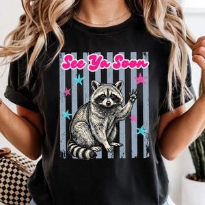 Peut inclure: T-shirt noir avec une illustration de raton laveur faisant un signe de la main. Le design comprend le texte "See Ya Soon" en rose, avec des &eacute;toiles bleues et roses et des rayures verticales grises. Le raton laveur est en noir et blanc.