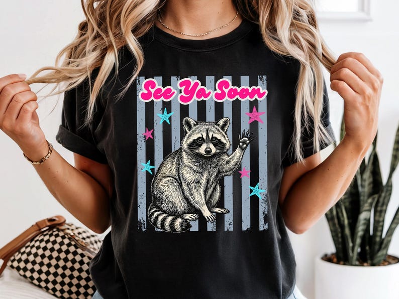 Peut inclure: T-shirt noir avec une illustration de raton laveur faisant un signe de la main. Le design comprend le texte "See Ya Soon" en rose, avec des &eacute;toiles bleues et roses et des rayures verticales grises. Le raton laveur est en noir et blanc.