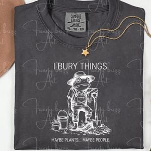 May include: Dark gray t-shirt with a white graphic of a frog in overalls holding a shovel, with the text "I BURY THINGS... MAYBE PLANTS... MAYBE PEOPLE." A gold star pendant necklace is draped on the shirt.