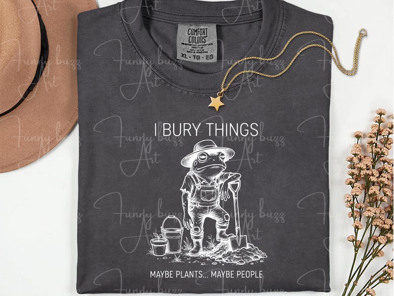 May include: Dark gray t-shirt with a white graphic of a frog in overalls holding a shovel, with the text "I BURY THINGS... MAYBE PLANTS... MAYBE PEOPLE." A gold star pendant necklace is draped on the shirt.
