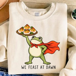 May include: A cream-coloured crewneck sweatshirt featuring a graphic of a green frog wearing a red cape and holding a platter of deviled eggs. Below the illustration, the text reads WE FEAST AT DAWN in black, capitalised, sans-serif lettering.