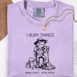 May include: A light purple t-shirt with the text "I BURY THINGS" and a graphic of a frog wearing overalls and a hat, holding a shovel. The text "MAYBE PLANTS... MAYBE PEOPLE" is below the frog. A gold star necklace is draped on the shirt.