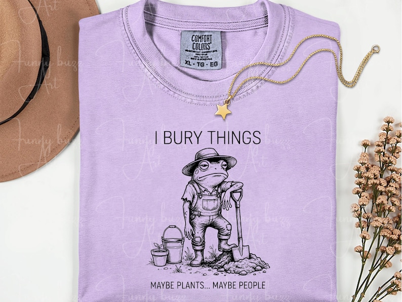 May include: A light purple t-shirt with the text "I BURY THINGS" and a graphic of a frog wearing overalls and a hat, holding a shovel. The text "MAYBE PLANTS... MAYBE PEOPLE" is below the frog. A gold star necklace is draped on the shirt.