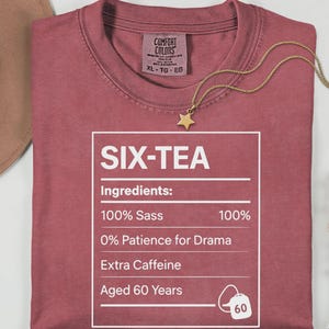 May include: A dusty rose t-shirt with the text "SIX-TEA" in a white box, listing ingredients like "100% Sass" and "Extra Caffeine." A gold star pendant necklace and a hat are next to the shirt. The shirt is XL-TG-EG.