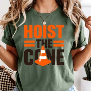 May include: Olive green t-shirt with the words "HOIST THE CONE" in orange and brown. The design includes an orange and white traffic cone graphic. The shirt is a casual, short-sleeved style.
