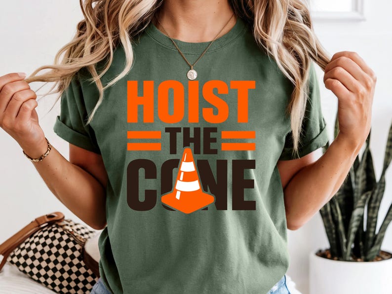 May include: Olive green t-shirt with the words "HOIST THE CONE" in orange and brown. The design includes an orange and white traffic cone graphic. The shirt is a casual, short-sleeved style.