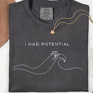 May include: Dark gray t-shirt with the text "I HAD POTENTIAL" and a white line drawing of a surfer on a wave. A gold star pendant hangs from a gold chain. A brown hat and pink flowers are in the background.