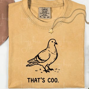 May include: Mustard-colored t-shirt with a black line drawing of a pigeon and the phrase "THAT'S COO." A gold-colored necklace with a star pendant is draped on the shirt. The shirt has a Comfort Colors tag.