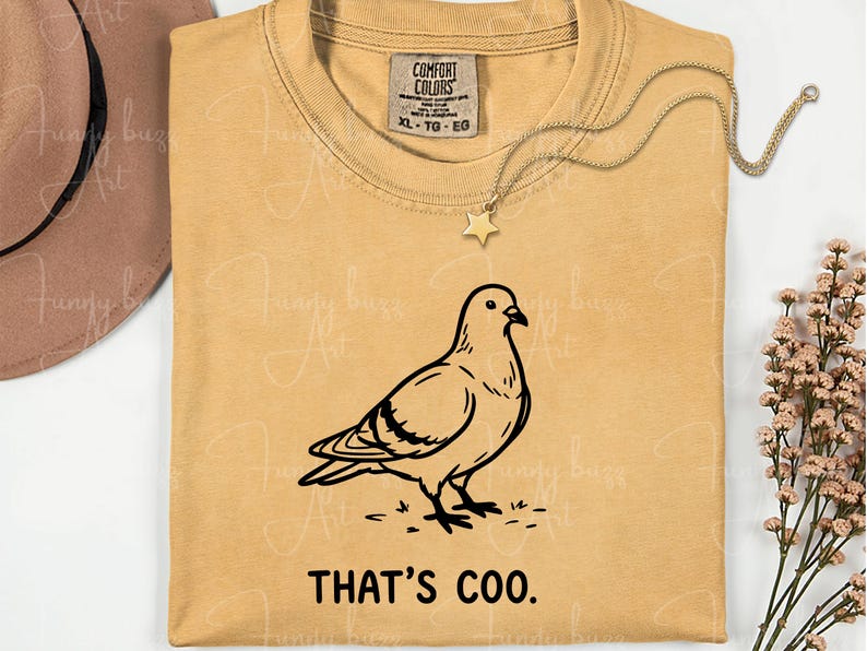 May include: Mustard-colored t-shirt with a black line drawing of a pigeon and the phrase "THAT'S COO." A gold-colored necklace with a star pendant is draped on the shirt. The shirt has a Comfort Colors tag.