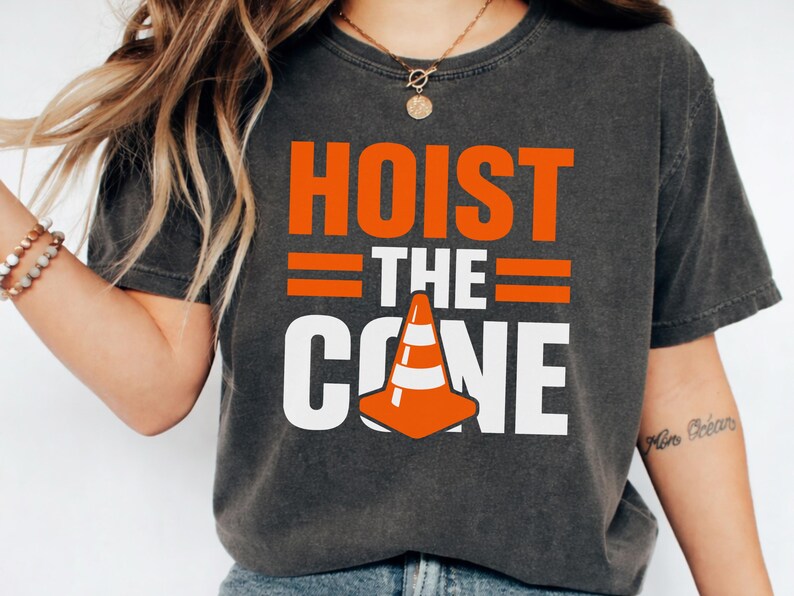 May include: Dark gray t-shirt with the words "HOIST THE CONE" in orange and white block letters. A graphic of an orange and white traffic cone is featured below the text. The shirt is a casual wear item.