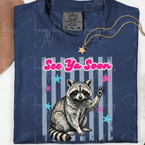 Peut inclure: T-shirt bleu marine avec un raton laveur qui salue et le texte "See Ya Soon". Le motif comprend des &eacute;toiles roses et bleues et des rayures grises. Un collier dor&eacute; avec un pendentif en forme d'&eacute;toile est pos&eacute; sur le t-shirt.