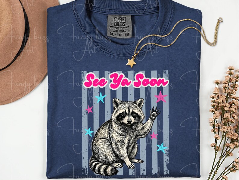Peut inclure: T-shirt bleu marine avec un raton laveur qui salue et le texte "See Ya Soon". Le motif comprend des &eacute;toiles roses et bleues et des rayures grises. Un collier dor&eacute; avec un pendentif en forme d'&eacute;toile est pos&eacute; sur le t-shirt.