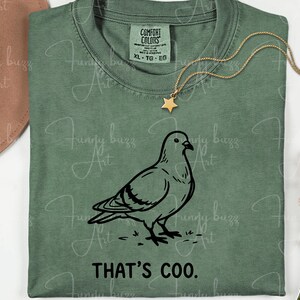 May include: Sage green t-shirt with a black graphic of a pigeon and the text "THAT'S COO." A gold necklace with a star pendant is draped over the shirt. The shirt is labeled XL-TG-EG.