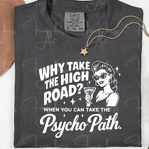 May include: Dark gray t-shirt with the text "WHY TAKE THE HIGH ROAD? WHEN YOU CAN TAKE THE Psycho Path." A graphic of a woman in sunglasses holding a martini glass is also on the shirt. A gold necklace with a star pendant is on the shirt.