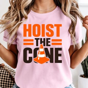 May include: Pink t-shirt with the words "HOIST THE CONE" in orange and black. The design includes a traffic cone graphic. The shirt is short-sleeved.