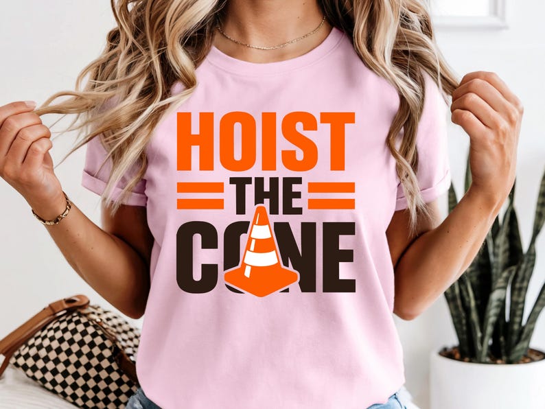 May include: Pink t-shirt with the words "HOIST THE CONE" in orange and black. The design includes a traffic cone graphic. The shirt is short-sleeved.