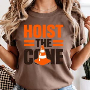 May include: A brown t-shirt with the words "HOIST THE CONE" in orange and black. The design includes a traffic cone graphic. The shirt is short-sleeved.