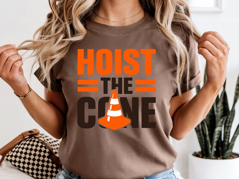 May include: A brown t-shirt with the words "HOIST THE CONE" in orange and black. The design includes a traffic cone graphic. The shirt is short-sleeved.