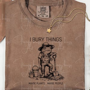 May include: Brown Comfort Colors t-shirt with a graphic of a frog in overalls holding a shovel, with the text "I BURY THINGS... MAYBE PLANTS... MAYBE PEOPLE." A gold star necklace and a hat are also visible.