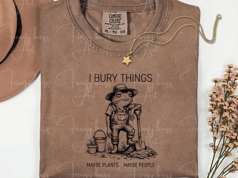 May include: Brown Comfort Colors t-shirt with a graphic of a frog in overalls holding a shovel, with the text "I BURY THINGS... MAYBE PLANTS... MAYBE PEOPLE." A gold star necklace and a hat are also visible.