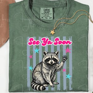 Peut inclure: T-shirt vert avec un dessin de raton laveur qui fait signe, le texte "See Ya Soon" et des &eacute;toiles. Un collier dor&eacute; avec un pendentif en forme d'&eacute;toile est pos&eacute; sur le t-shirt. Taille XL-TG-EG.
