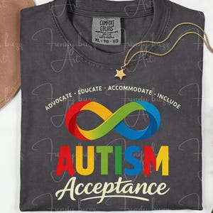 Puede incluir: Camiseta gris oscuro con las palabras "AUTISM Acceptance" en letras coloridas y un símbolo de infinito arcoíris. La camiseta también tiene las palabras "ADVOCATE EDUCATE ACCOMMODATE INCLUDE". Un collar de estrella dorada está sobre la camiseta.