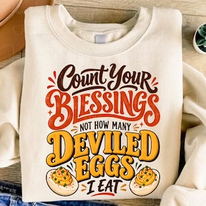 May include: A cream-coloured crewneck sweatshirt featuring a graphic print with the text "Count Your BLESSINGS NOT HOW MANY DEVILED EGGS I EAT" in brown, orange, and yellow, accompanied by illustrations of two halved deviled eggs with green garnish.