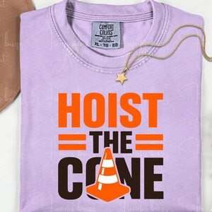 May include: A light purple t-shirt with the words "HOIST THE CONE" in orange and black. The design includes a traffic cone graphic. A gold star pendant necklace and a brown hat are also visible.