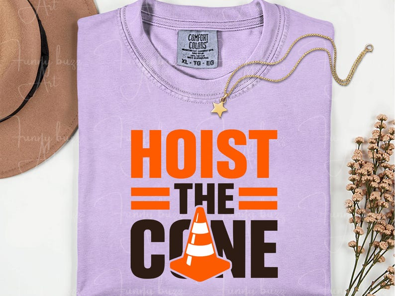 May include: A light purple t-shirt with the words "HOIST THE CONE" in orange and black. The design includes a traffic cone graphic. A gold star pendant necklace and a brown hat are also visible.