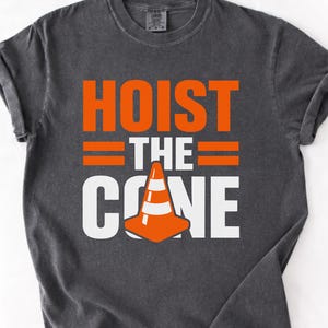 May include: Dark gray t-shirt with the words "HOIST THE CONE" in orange and white block letters. The design includes a traffic cone graphic. The shirt has rolled-up sleeves.