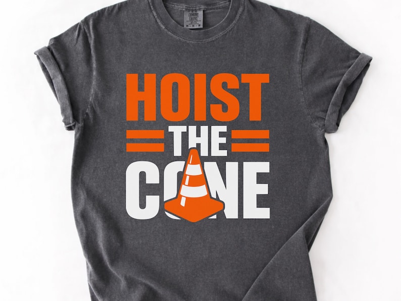 May include: Dark gray t-shirt with the words "HOIST THE CONE" in orange and white block letters. The design includes a traffic cone graphic. The shirt has rolled-up sleeves.