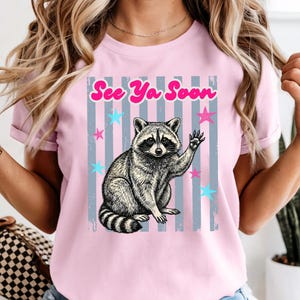 Peut inclure: T-shirt rose clair avec une illustration de raton laveur qui salue. Le design comprend le texte "See Ya Soon" en rose, avec des &eacute;toiles bleues et roses et des rayures verticales grises. Un t-shirt graphique d&eacute;contract&eacute; et amusant.