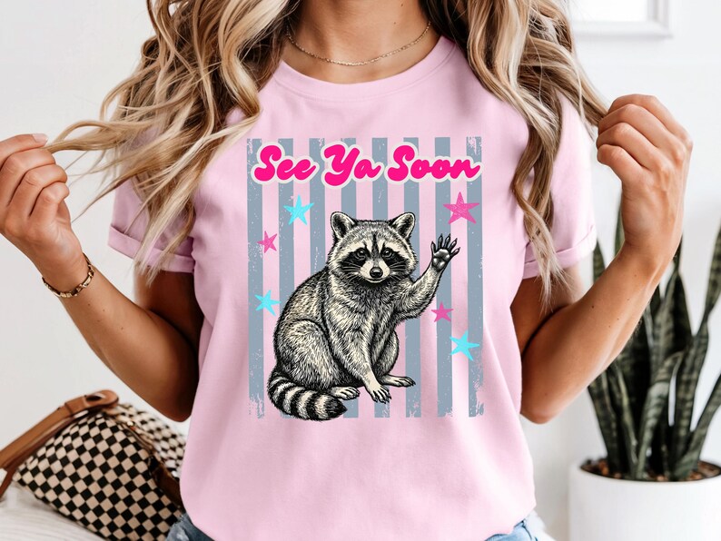 Peut inclure: T-shirt rose clair avec une illustration de raton laveur qui salue. Le design comprend le texte "See Ya Soon" en rose, avec des &eacute;toiles bleues et roses et des rayures verticales grises. Un t-shirt graphique d&eacute;contract&eacute; et amusant.