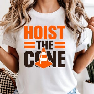 May include: White t-shirt with the text "HOIST THE CONE" in orange and black. The design includes a traffic cone graphic. The shirt is a crew neck style.