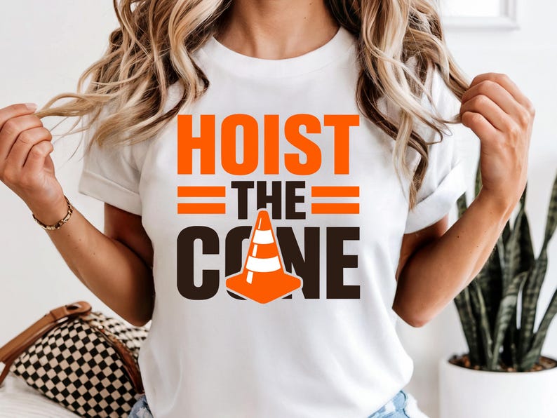 May include: White t-shirt with the text "HOIST THE CONE" in orange and black. The design includes a traffic cone graphic. The shirt is a crew neck style.