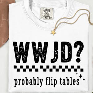 May include: White t-shirt with the text "WWJD? probably flip tables" in black. The shirt has a crew neck and a tag that says "Comfort Colors". A gold star pendant necklace is draped on the shirt.