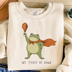 Op de afbeelding: Een crèmekleurige sweater met ronde hals en een afbeelding van een groene kikker met een oranje cape die een gefrituurde kippenpoot vasthoudt. Onder de illustratie staat de tekst WE FEAST AT DAWN in een eenvoudig, donker lettertype.