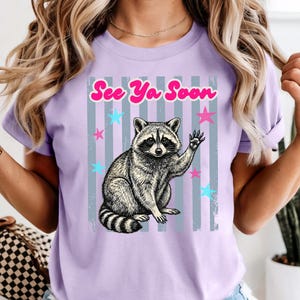 Peut inclure: T-shirt lilas avec une illustration de raton laveur qui fait signe. Le design comprend le texte "See Ya Soon" en rose, avec des &eacute;toiles bleues et roses et des rayures grises. Le raton laveur est en noir et blanc.