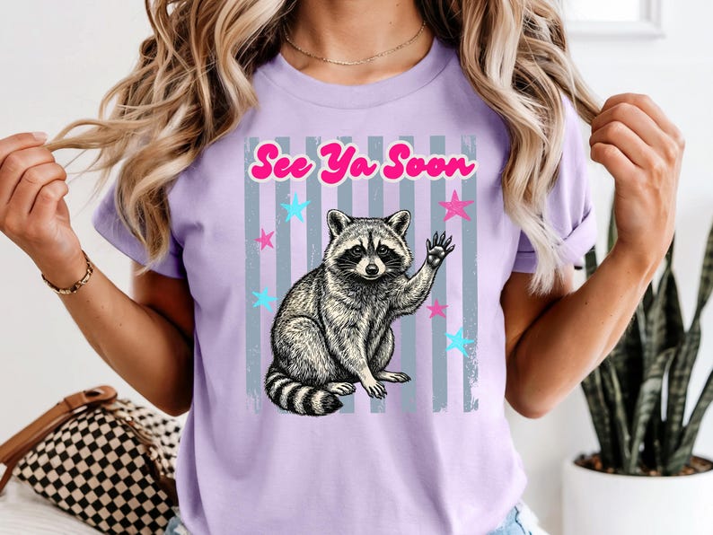 Peut inclure: T-shirt lilas avec une illustration de raton laveur qui fait signe. Le design comprend le texte "See Ya Soon" en rose, avec des &eacute;toiles bleues et roses et des rayures grises. Le raton laveur est en noir et blanc.