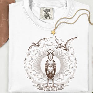 Puede incluir: Camiseta blanca con una ilustración marrón de un picozapato rodeado de pterodáctilos y un diseño de rayos de sol. También se ven un collar con colgante de estrella dorada y un sombrero marrón.