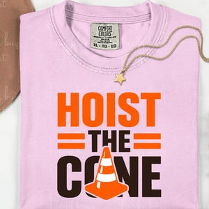 May include: A light pink t-shirt with the words "HOIST THE CONE" in orange and black, featuring a traffic cone graphic. A gold necklace with a star pendant rests on the shirt.