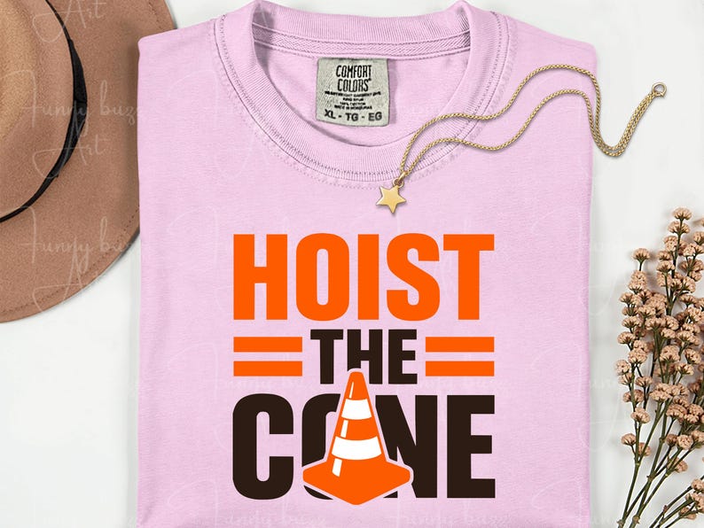 May include: A light pink t-shirt with the words "HOIST THE CONE" in orange and black, featuring a traffic cone graphic. A gold necklace with a star pendant rests on the shirt.