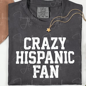 Puede incluir: Camiseta gris oscuro con el texto blanco "CRAZY HISPANIC FAN". También son visibles un collar con colgante de estrella dorada y un sombrero marrón. La camiseta tiene una etiqueta Comfort Colors.