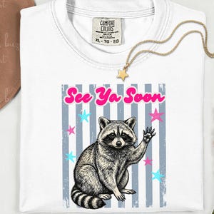 Peut inclure: T-shirt blanc avec un dessin de raton laveur qui salue, le texte "See Ya Soon" et des &eacute;toiles. Le t-shirt a un col rond et une &eacute;tiquette "Comfort Colors". Un collier &eacute;toile dor&eacute; est pos&eacute; sur le t-shirt.