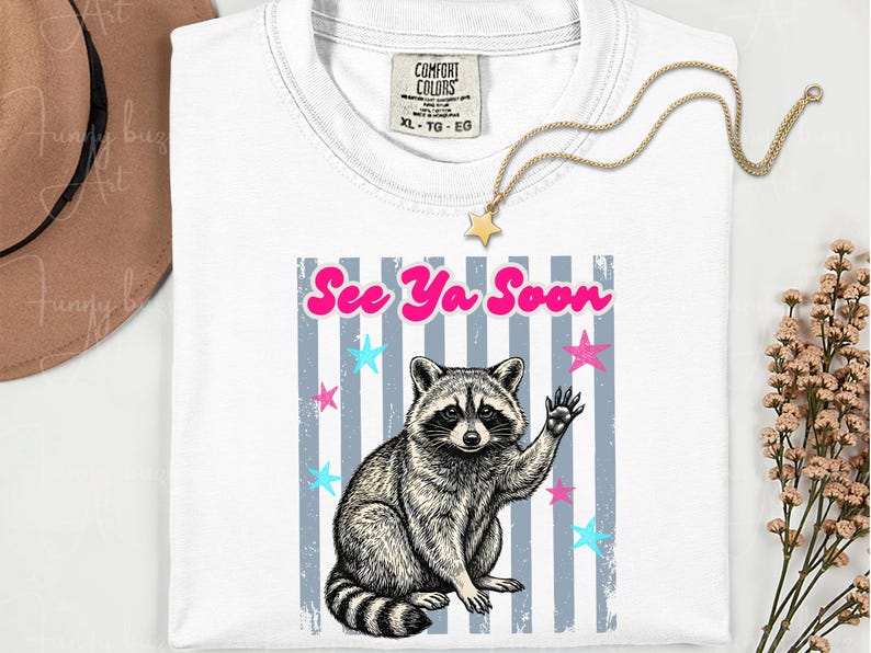 Peut inclure: T-shirt blanc avec un dessin de raton laveur qui salue, le texte "See Ya Soon" et des &eacute;toiles. Le t-shirt a un col rond et une &eacute;tiquette "Comfort Colors". Un collier &eacute;toile dor&eacute; est pos&eacute; sur le t-shirt.