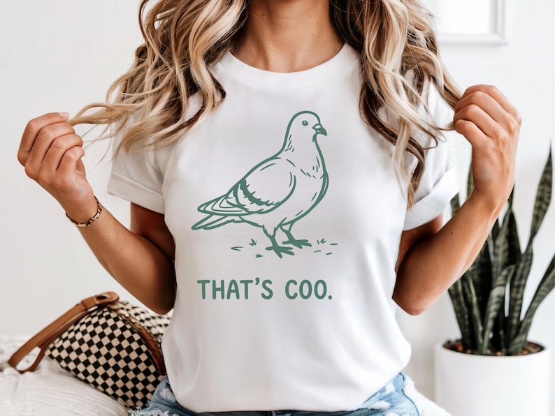 May include: White t-shirt featuring a green illustration of a pigeon and the phrase "THAT'S COO." The design is simple and clean, with the bird standing on a patch of grass. The shirt is a casual, everyday wear item.