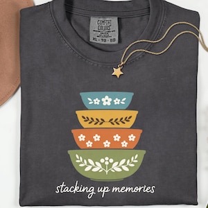 May include: Dark gray t-shirt with a stack of four decorative bowls in blue, yellow, orange, and green. Each bowl has a unique floral design. The text "stacking up memories" is printed below the bowls. A gold star necklace is draped on the shirt.