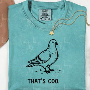 May include: A turquoise Comfort Colors t-shirt featuring a black line drawing of a pigeon and the phrase "THAT'S COO." A gold star pendant necklace rests on the shirt. The shirt is size XL.