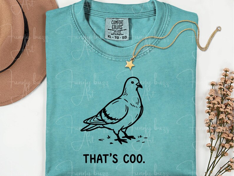 May include: A turquoise Comfort Colors t-shirt featuring a black line drawing of a pigeon and the phrase "THAT'S COO." A gold star pendant necklace rests on the shirt. The shirt is size XL.
