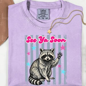 Peut inclure: T-shirt lilas avec un dessin de raton laveur qui salue et le texte "See Ya Soon". Le motif comprend des &eacute;toiles roses et bleues et des rayures verticales grises. Un collier dor&eacute; avec un pendentif en forme d'&eacute;toile est pos&eacute; sur le t-shirt.